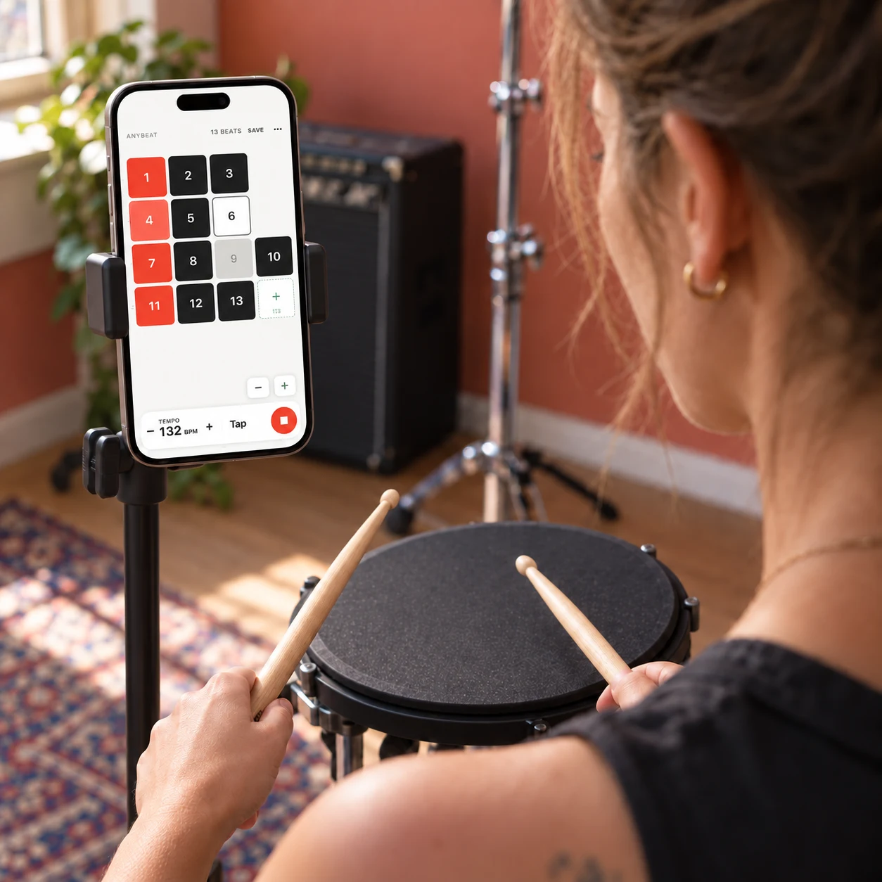 Over-the-shoulder scene of a woman practicing with two drumsticks while viewing Anybeat's rhythm grid and tempo controls on an iPhone mounted beside her practice pad